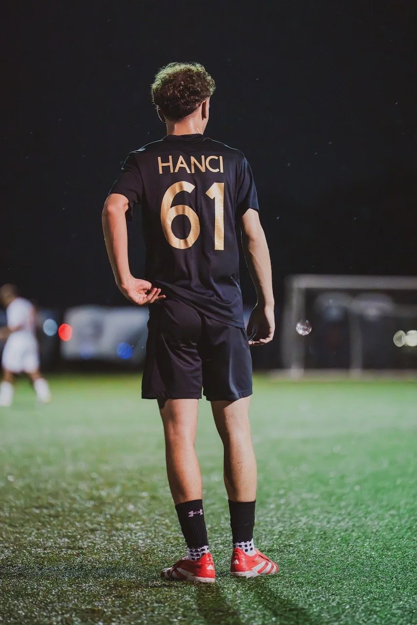 A soccer player wearing a black uniform with the name 'HANCI' and the number 61 on the back, standing on a field at night.