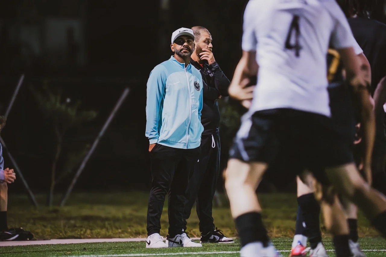Two soccer coaches standing on the sideline during a game at night, with players in white and black uniforms on the field in the foreground.