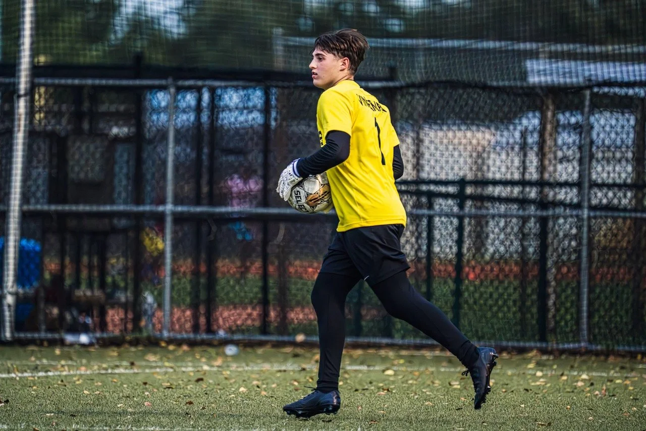 A soccer goalkeeper in a yellow jersey and black shorts holding a soccer ball on a field with a fenced background.