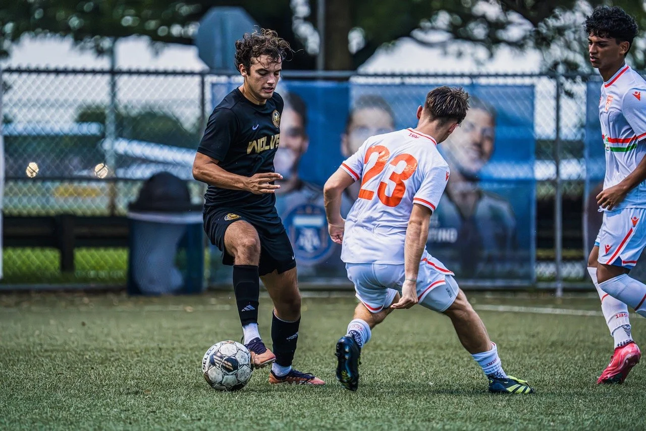 Two soccer players competing for the ball on a grass field during a match. One player in a black uniform and the other in a white uniform with red and blue accents. A fence and trees are visible in the background.