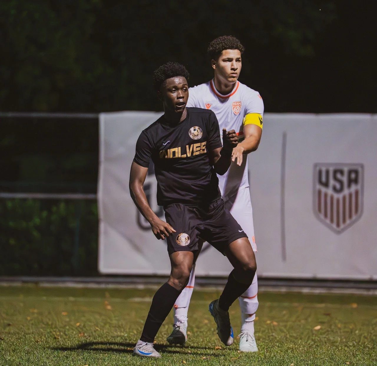 Two soccer players, one in a black uniform and the other in a white uniform with a yellow captain's armband, playing on a field at night during a game.