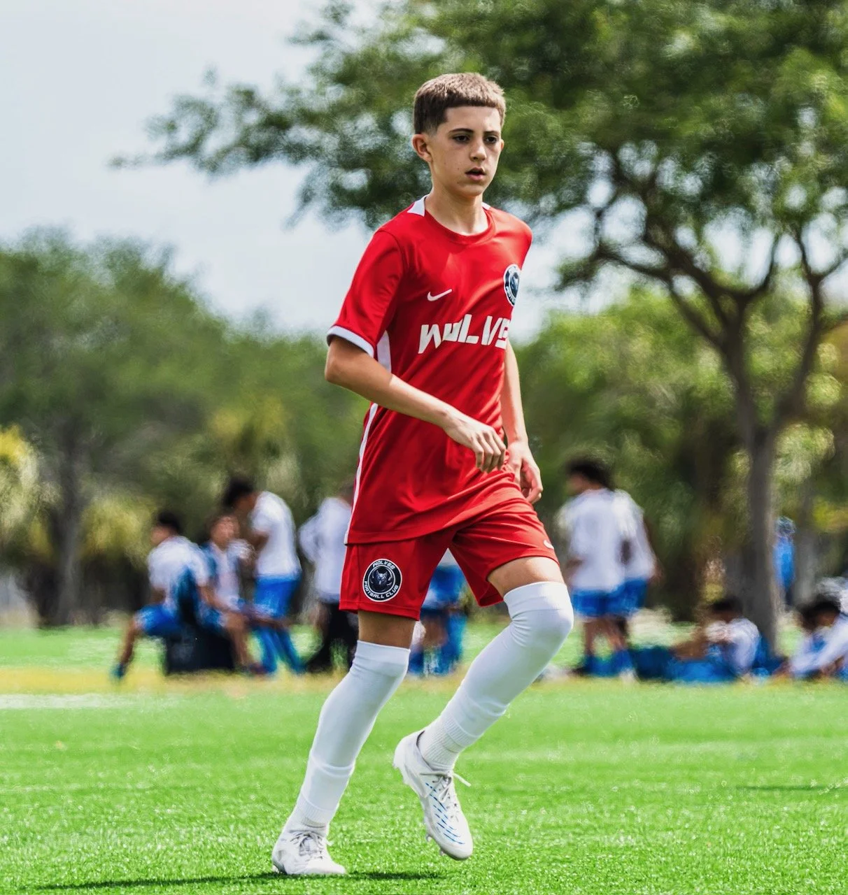 A young male soccer player in a red uniform with white socks and cleats playing on a grassy field during daytime, with trees and other players in the background.