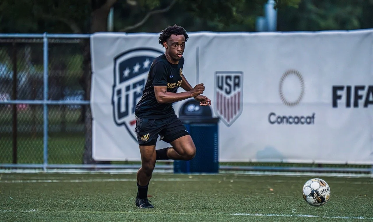 A man playing soccer on a field, wearing a black uniform with US Soccer logos, running toward the ball.