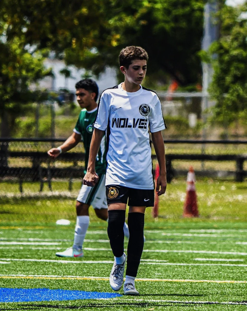 A boy in a white soccer jersey and black shorts walking on a soccer field, with another boy in a green jersey in the background.