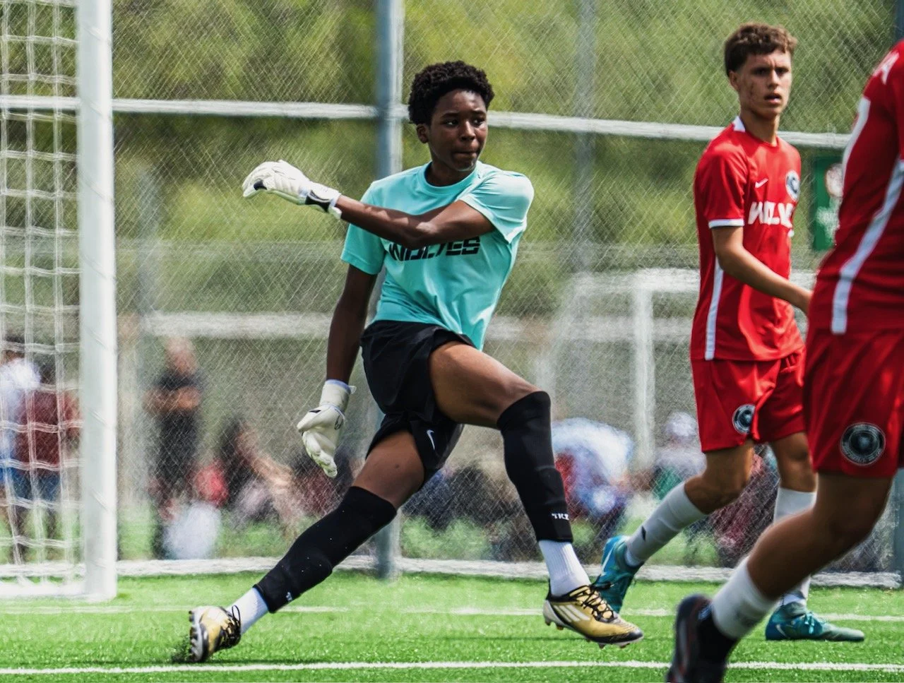 A young soccer goalkeeper in a teal jersey and black shorts making a save during a match on a green field, with other players in red jerseys nearby.