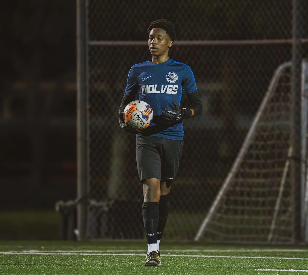 A young male goalkeeper holding a soccer ball on a soccer field at night, wearing a blue jersey and black shorts.
