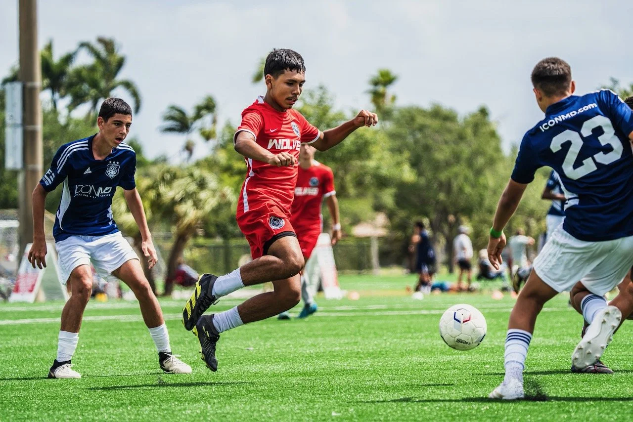 Young soccer players competing for the ball on the field during a game, with trees and spectators in the background.
