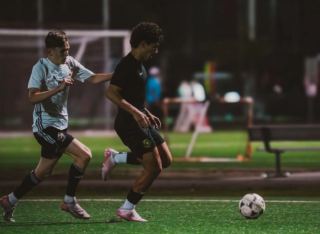 Two young soccer players chase a ball on a field during nighttime practice. One wears a white jersey and the other a black jersey.
