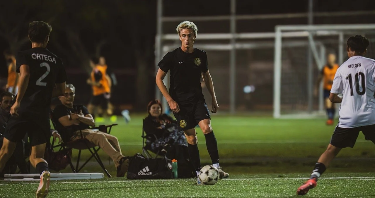 A soccer player with blonde hair in black uniform preparing to kick a soccer ball on the field during a game at night.