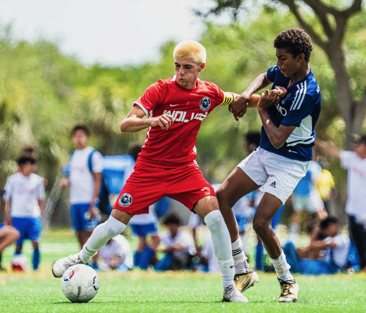 Two young soccer players contest for the ball during a match, with spectators blurred in the background.