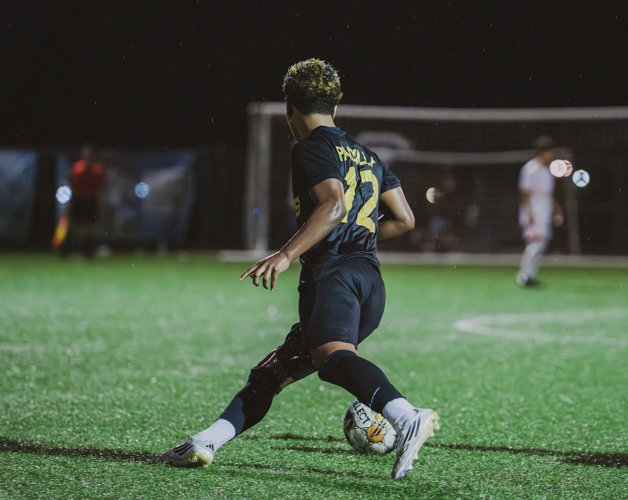 A soccer player in a black uniform kicking a ball on a wet, illuminated field at night.