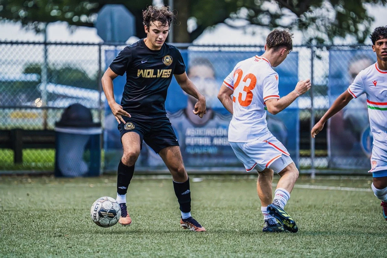 Young boys playing soccer on a field, with a fence and trees in the background. One boy is in a black uniform controlling the ball while two others in white uniforms run nearby.
