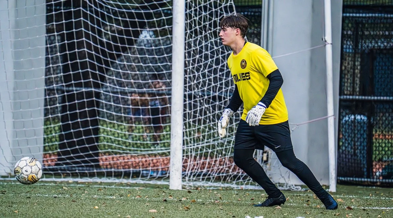 A young male soccer goalkeeper wearing a yellow jersey, black pants, and gloves, standing in front of a goal, preparing to stop a soccer ball on the field.