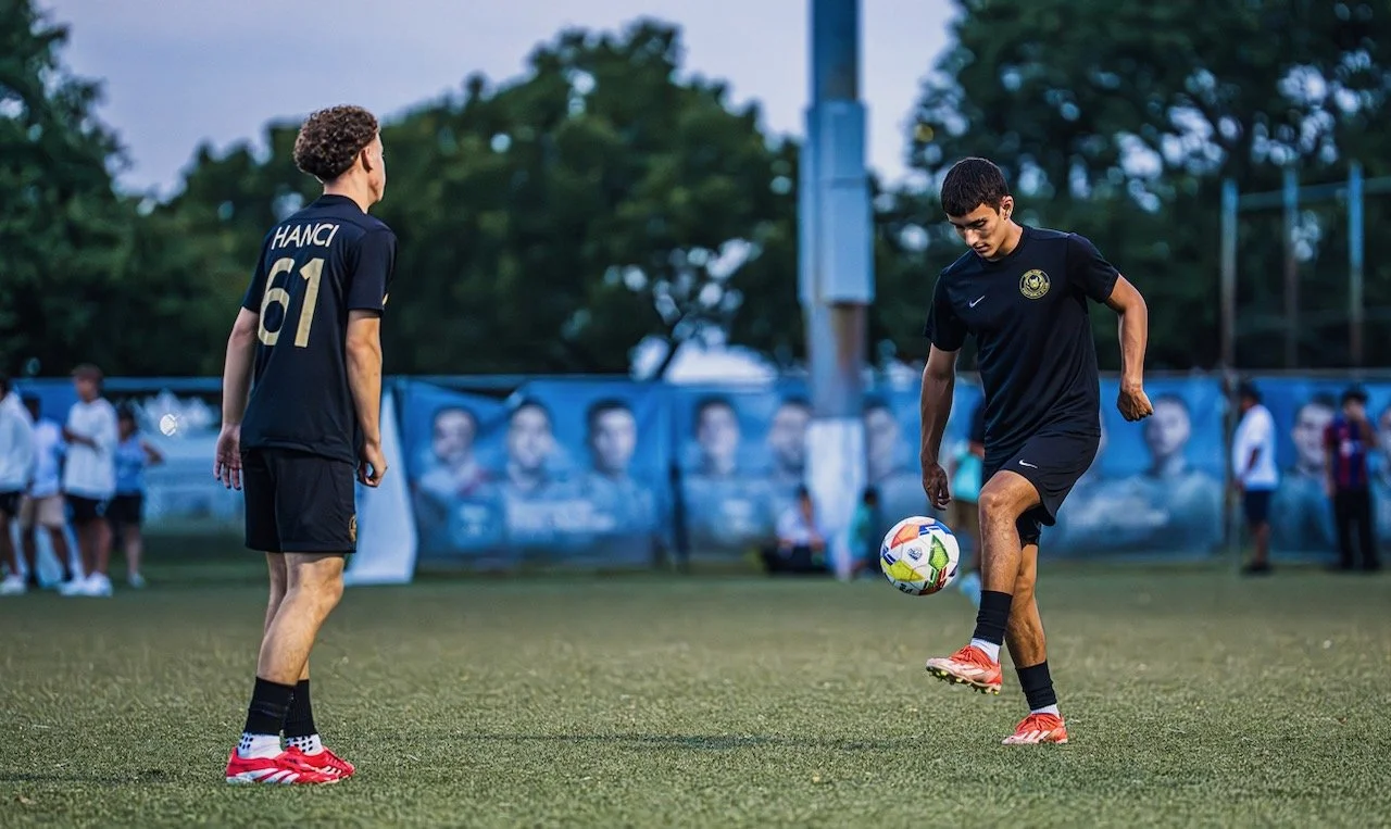 Two young soccer players in black uniforms on the field; one is juggling a soccer ball, and the other is observing. Several people are in the background.