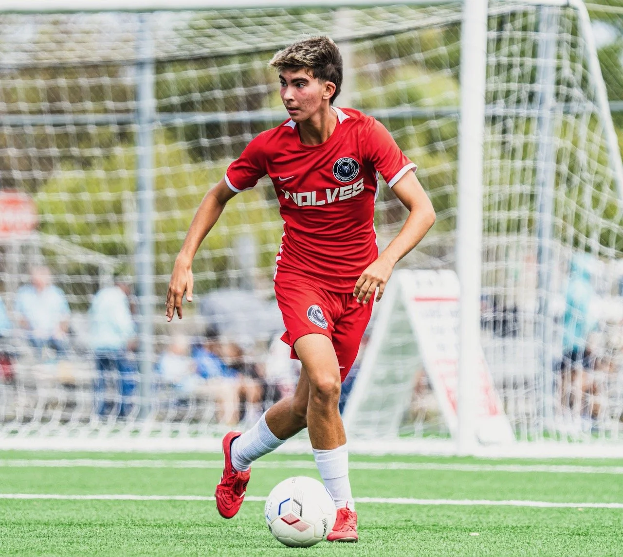 A young male soccer player in a red uniform dribbling a white soccer ball on an outdoor field with a goal in the background.