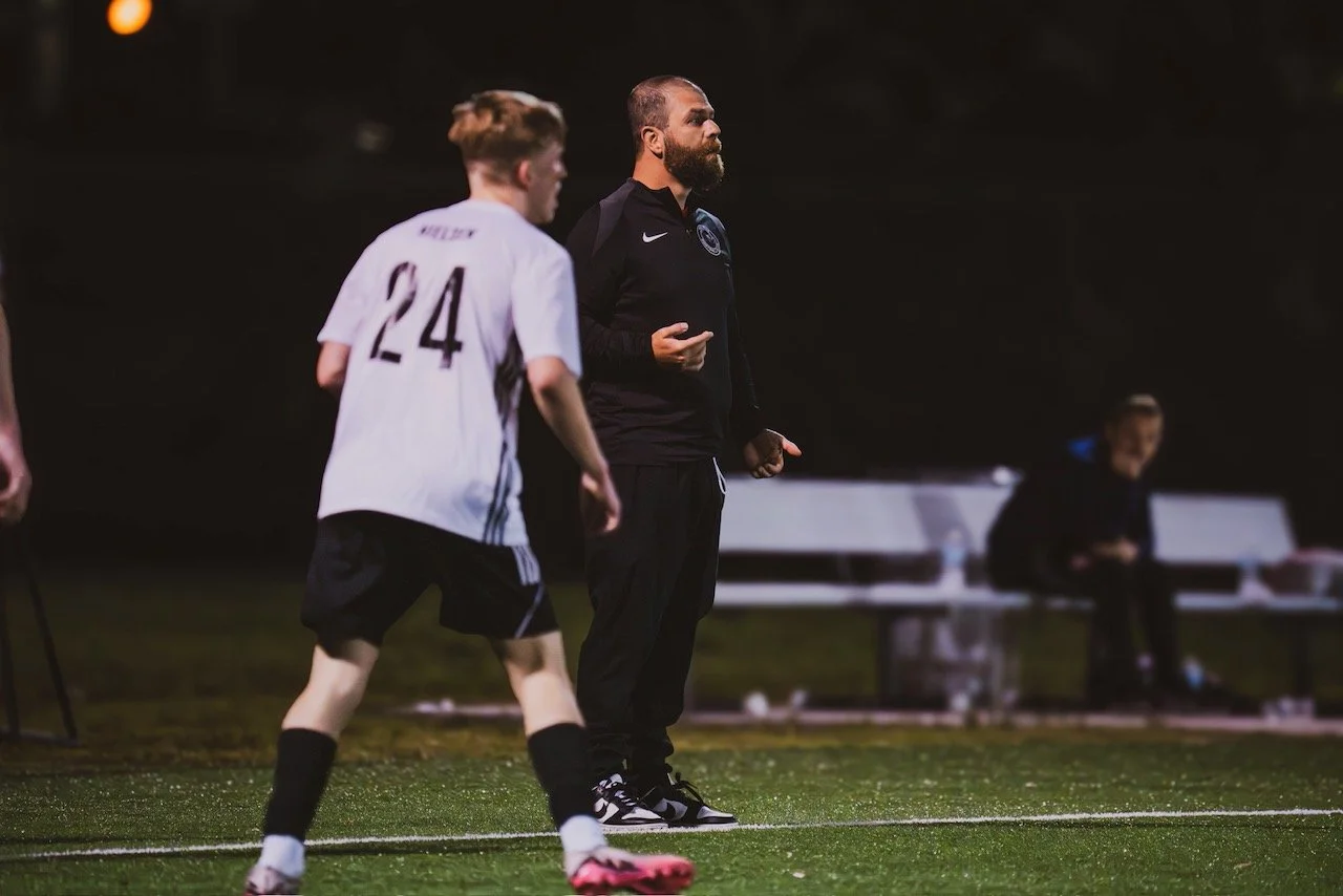 A soccer coach standing on the sideline of a field during a night game, watching players with a bench and a person sitting in the background.
