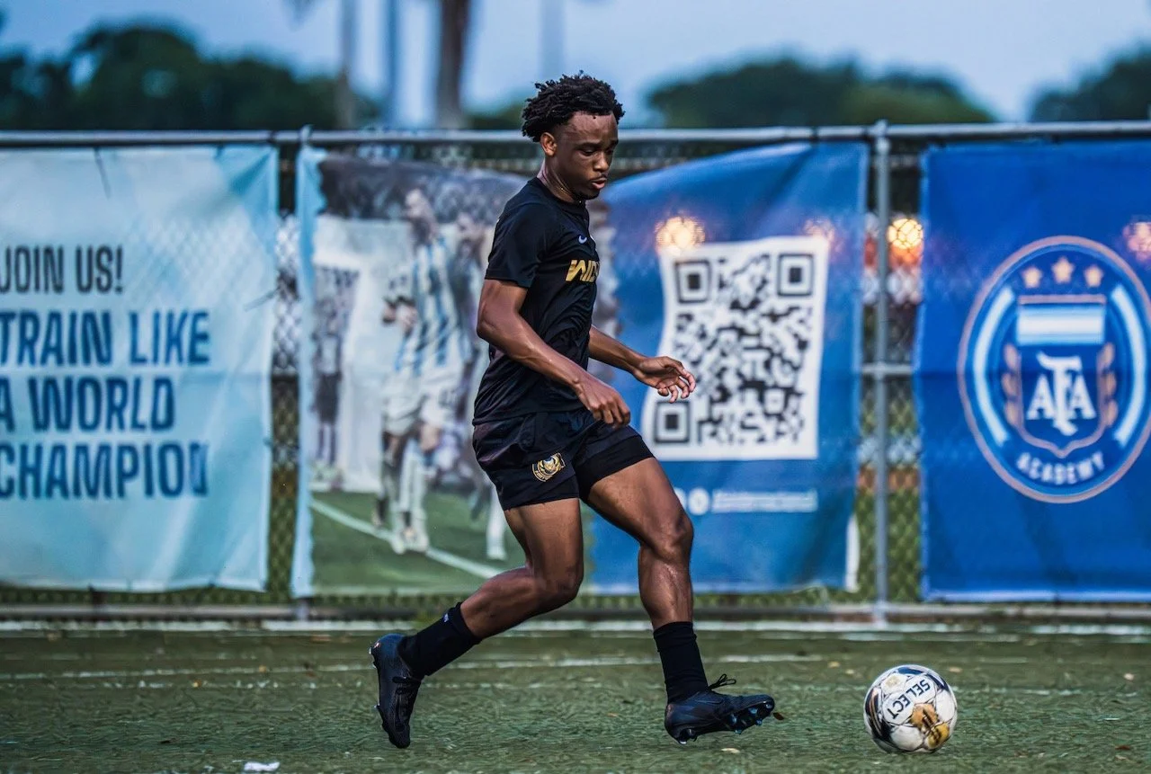A soccer player in black uniform practicing on a field with banners in the background, including a QR code and the AFA Academy logo.