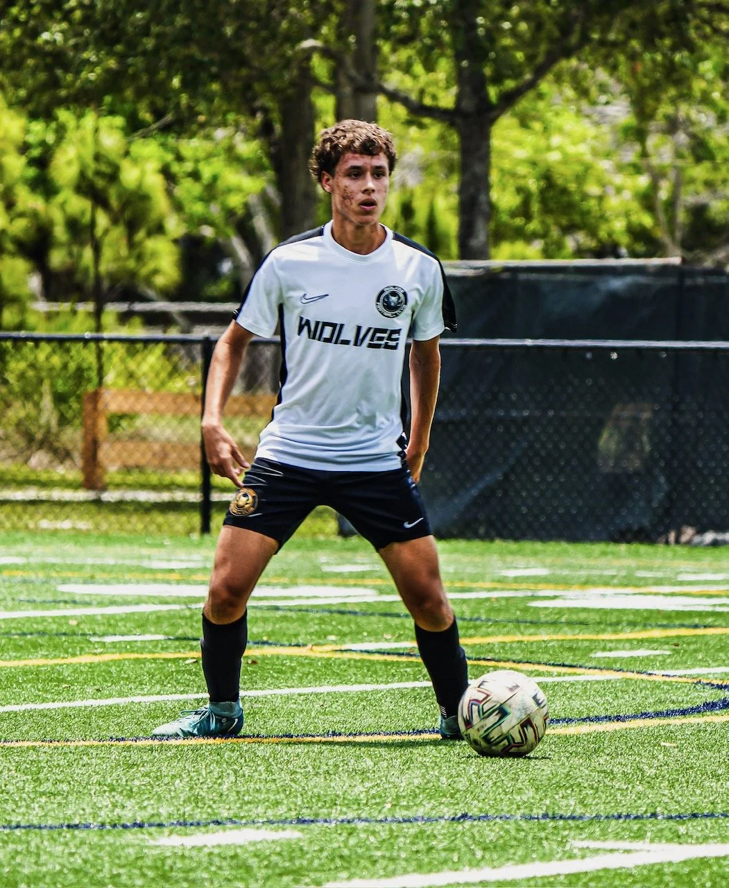 A young man in a white and black soccer uniform standing on a green soccer field with a ball at his feet, surrounded by trees and a black fence.