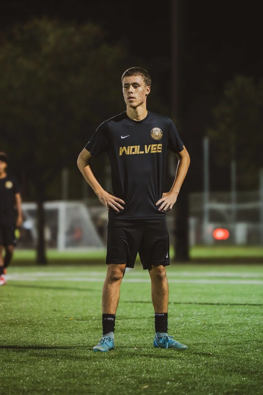 A young male soccer player standing on a soccer field at night, wearing a black uniform with 'WOLVES' written on the front, blue cleats, and black socks.