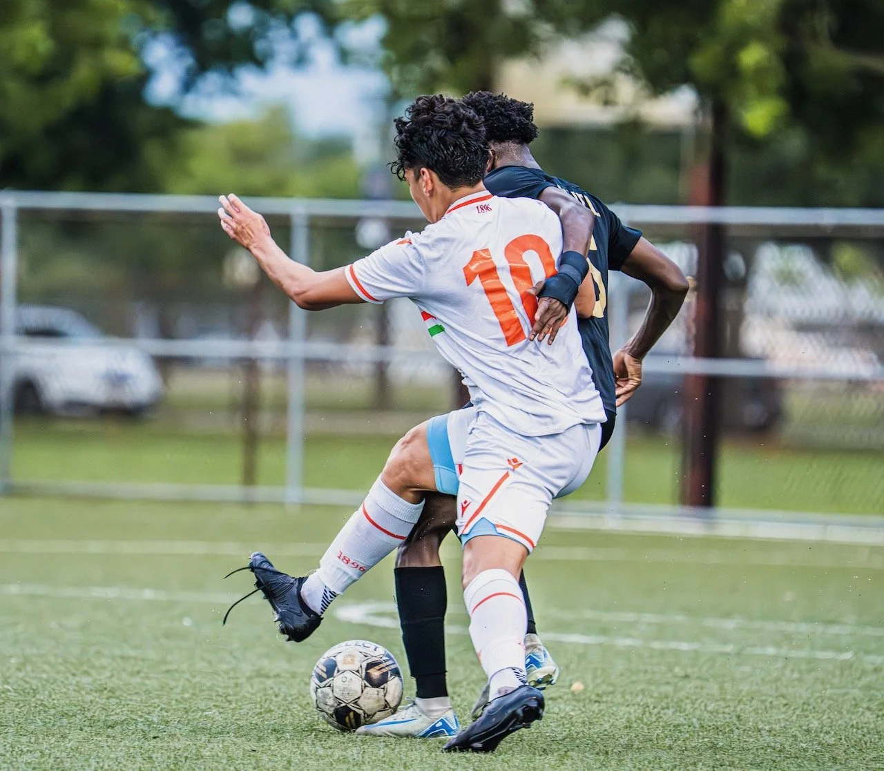 Two soccer players in a struggle for the ball on a field, one in a white jersey with the number 10, the other in a black jersey, with a fence and trees in the background.