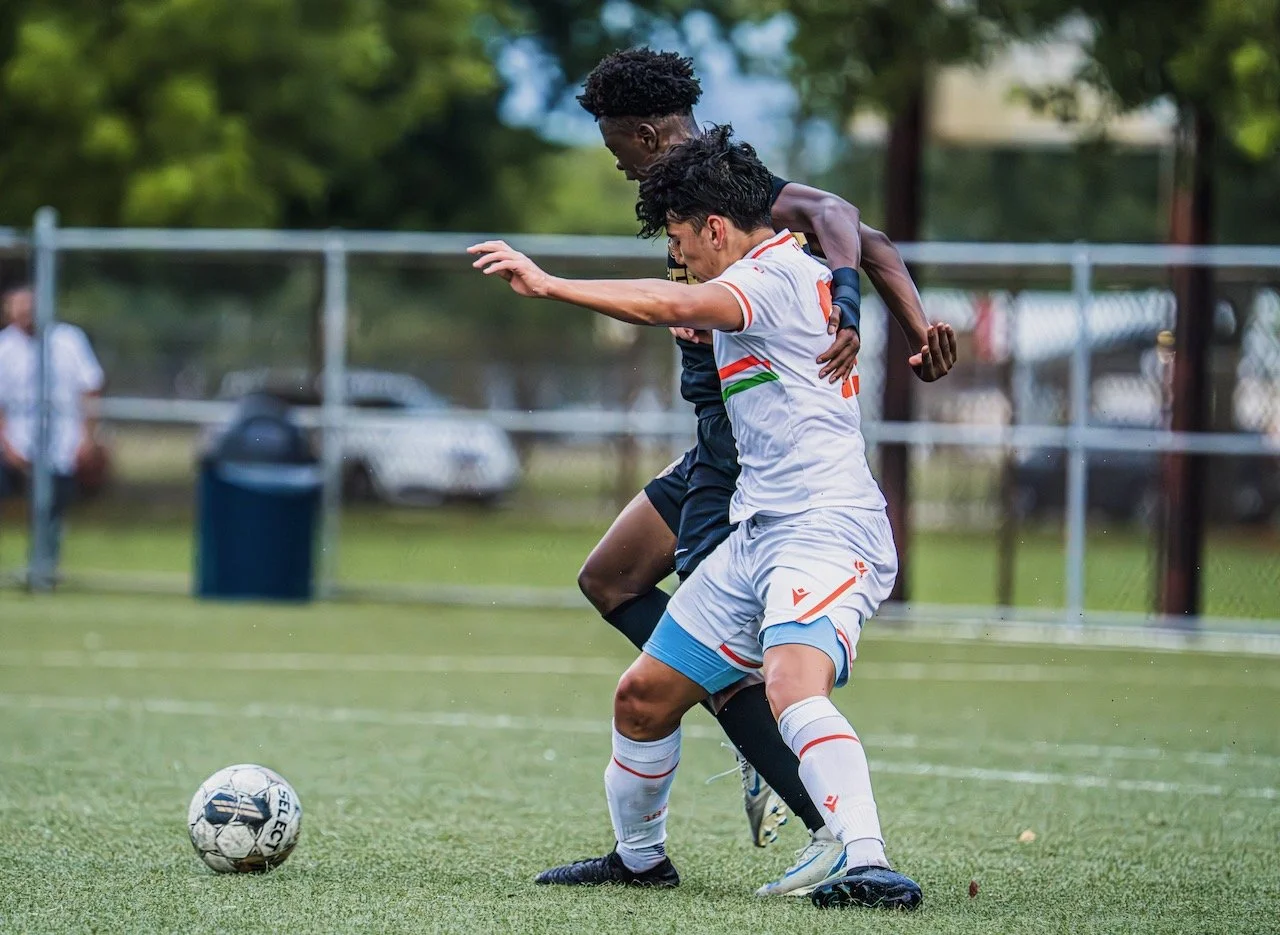 Two soccer players compete for the ball on a field during a game, with a green outdoor setting and a fence in the background.