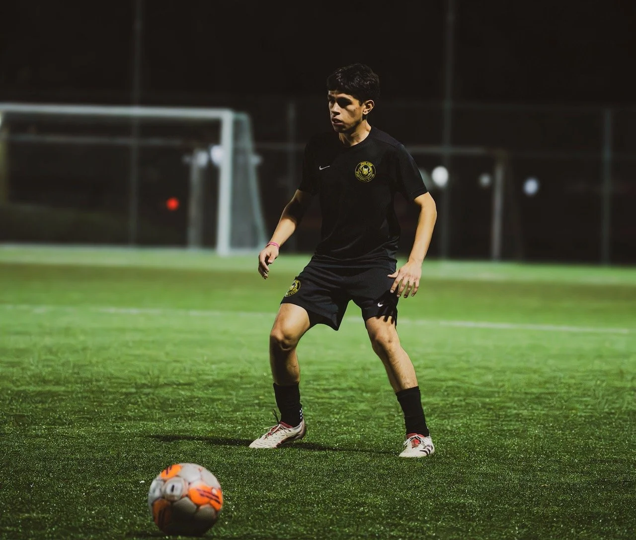 A young man playing soccer on a field at night, wearing a black sports uniform and preparing to kick a soccer ball.