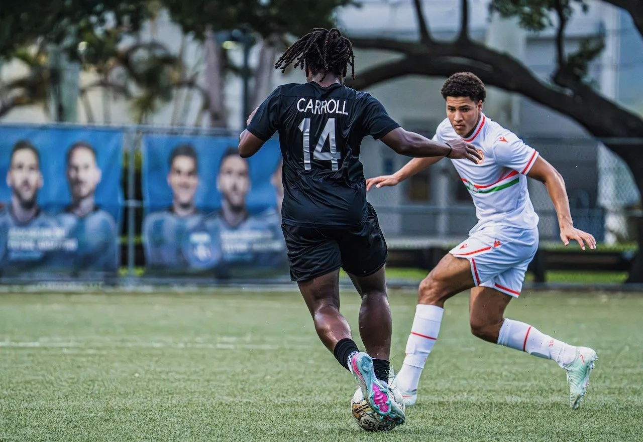 Two soccer players competing for the ball on the field, one in black with 'Carroll 14' on the jersey, the other in white with red and green accents, during a match with advertisements in the background.