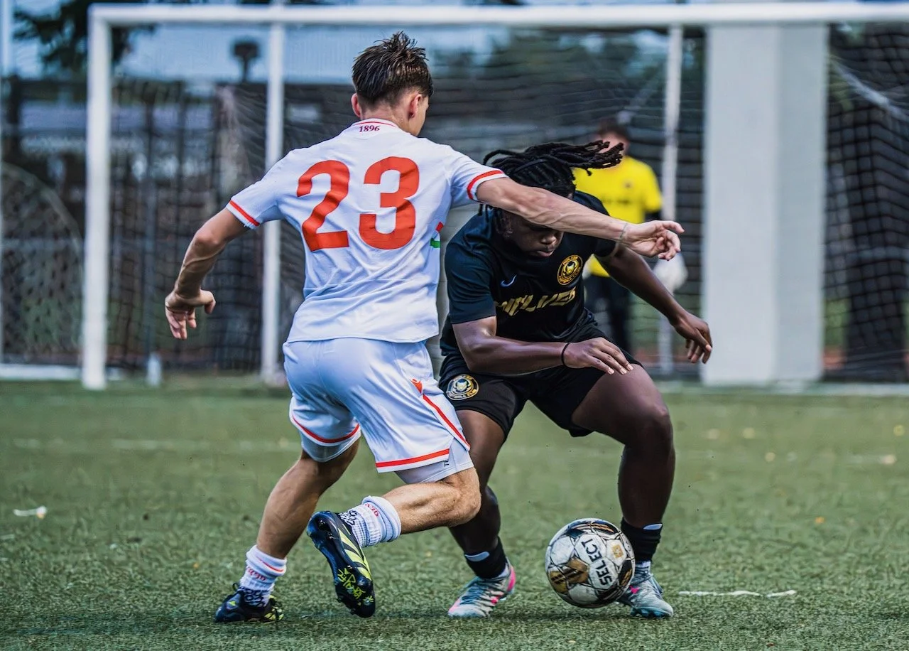 Two soccer players competing for the ball during a match on an outdoor field, with a goalkeeper in the background.