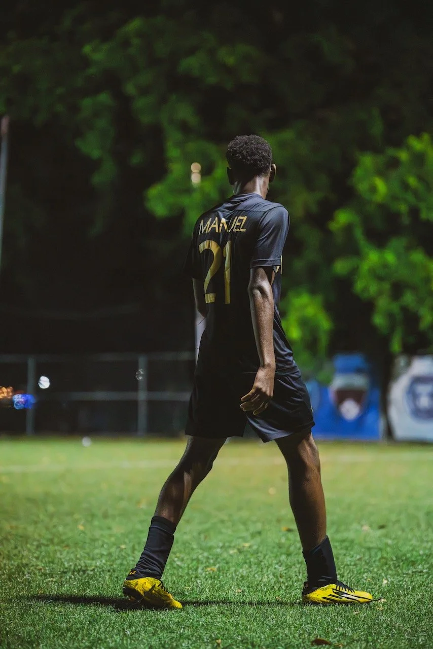 A young male soccer player wearing a black uniform with yellow accents, standing on a grassy field at night, with his back facing the camera. The uniform has the name 'MANEL' and the number '21' printed on it. The background is dark with some blue an