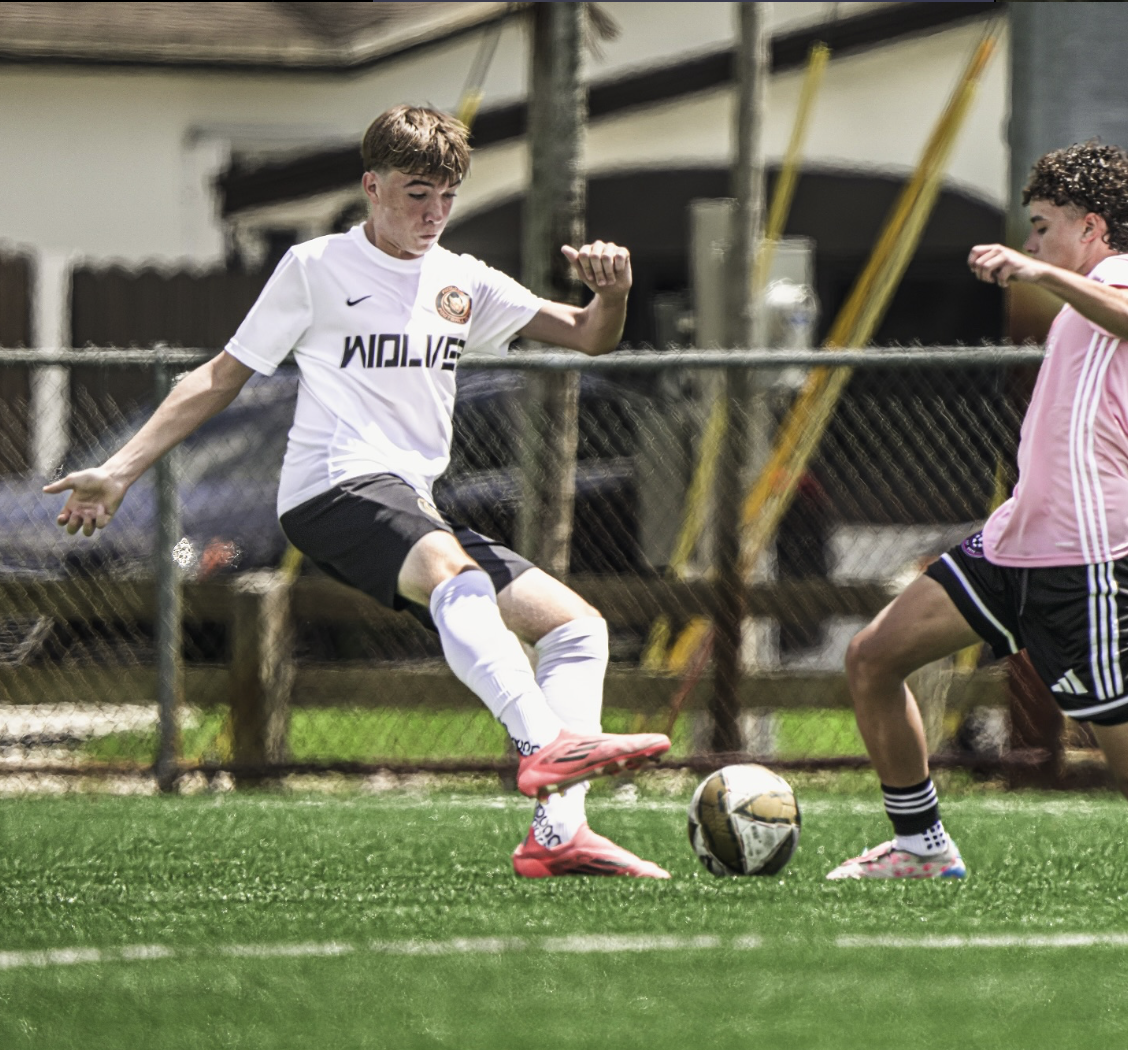 Two young boys playing soccer outdoors; one in a white jersey kicking the ball, the other in a pink jersey defending; grassy field and fence in background.