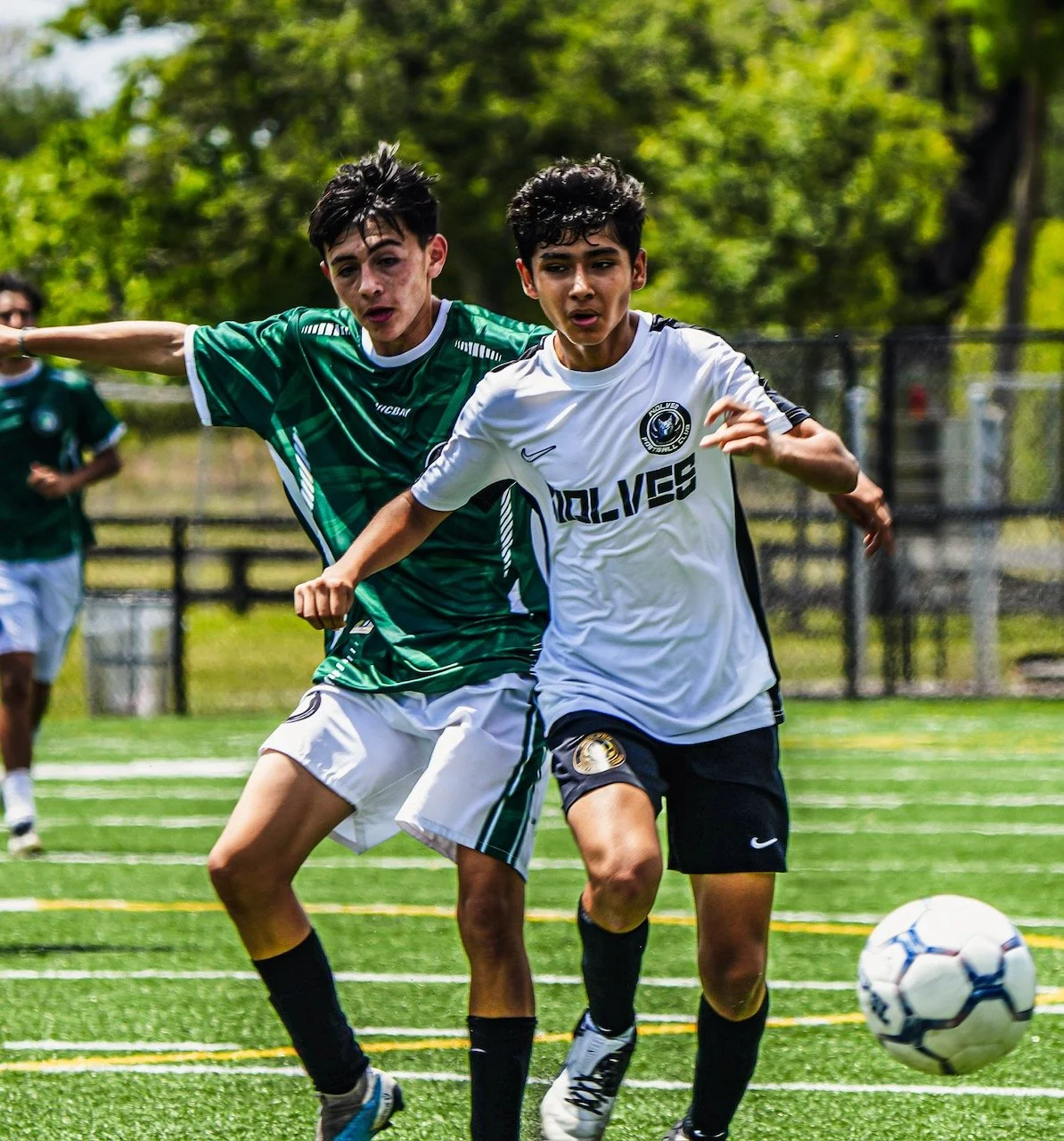 Two young male soccer players competing for the ball on a sunny field, with one in a green jersey and the other in a white jersey, during a match.
