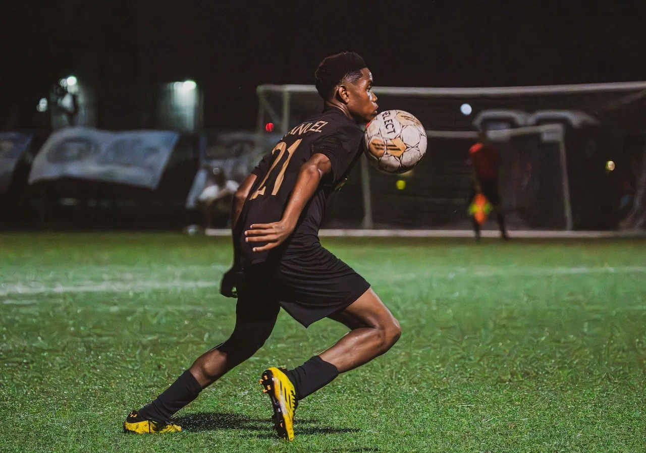 A soccer player in black uniform is about to kick a soccer ball during a nighttime game on a grassy field.