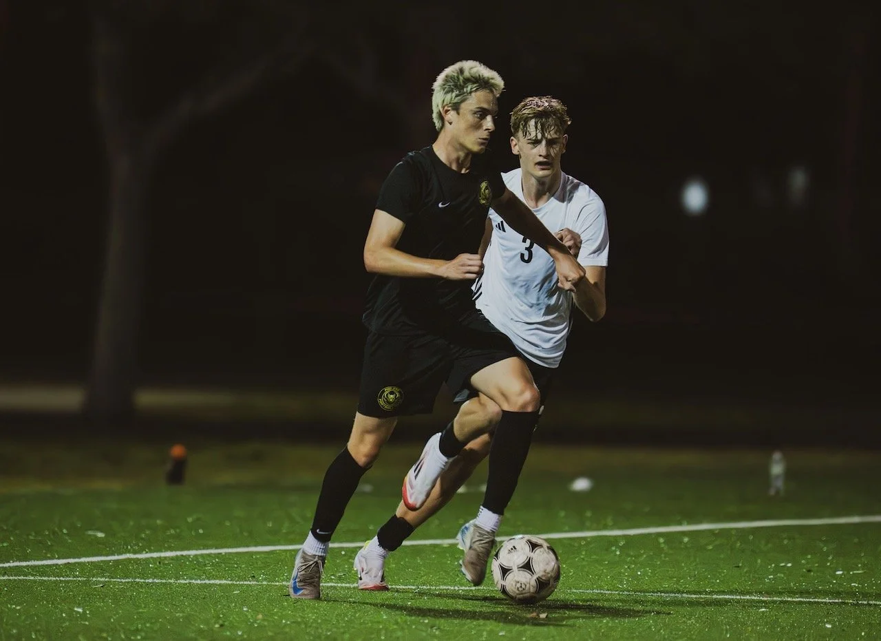 Two soccer players, one in a black uniform and the other in a white uniform, competing for the ball on a green field at night.