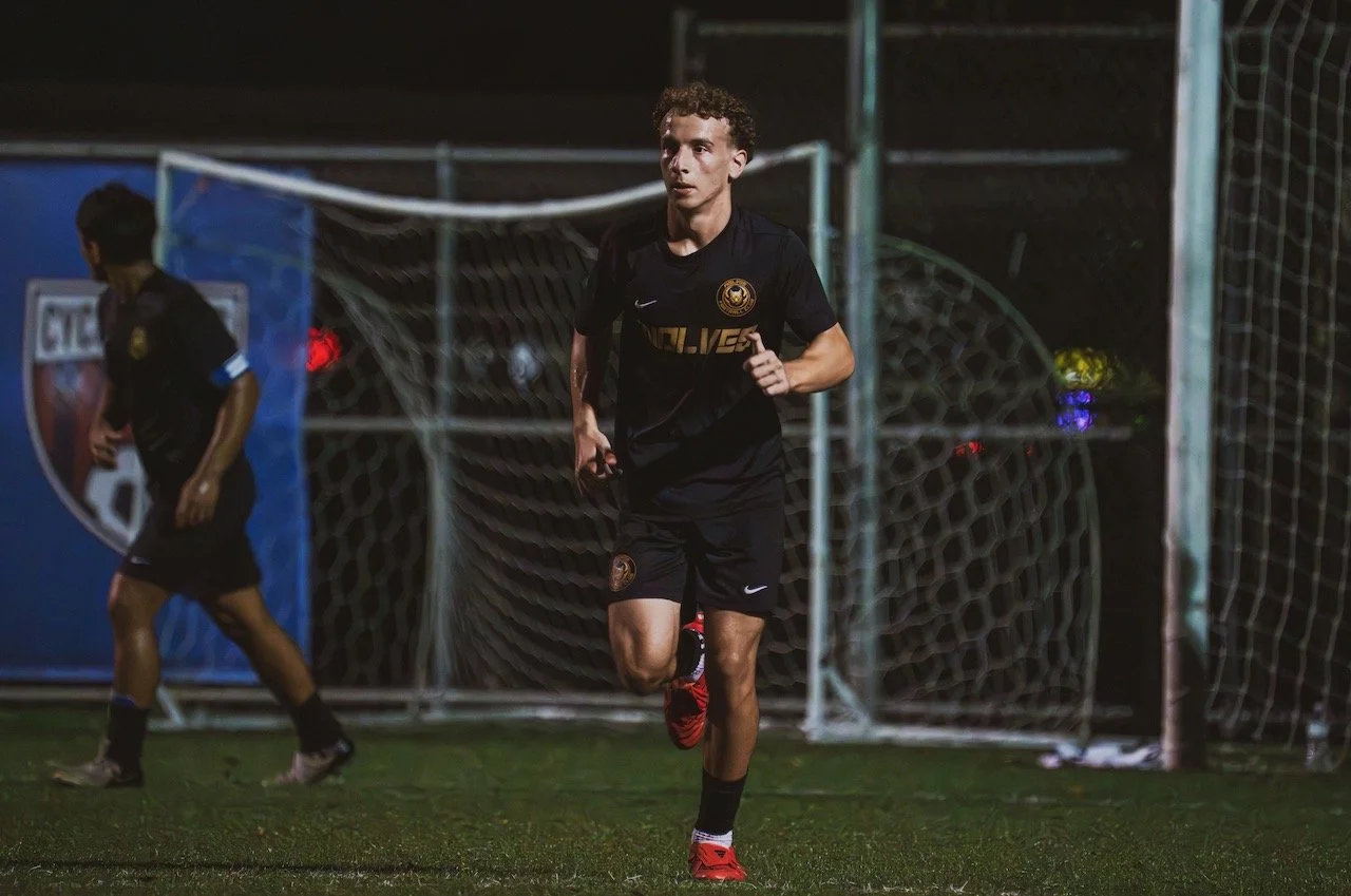 A soccer player running on the field at night, wearing a black uniform, near the goal with netting.