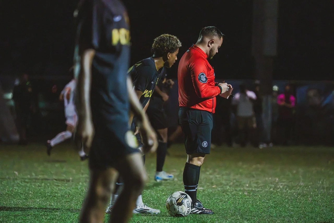 A soccer referee with a notepad standing on the field with a soccer ball at his feet during a night game, with players and spectators in the background.