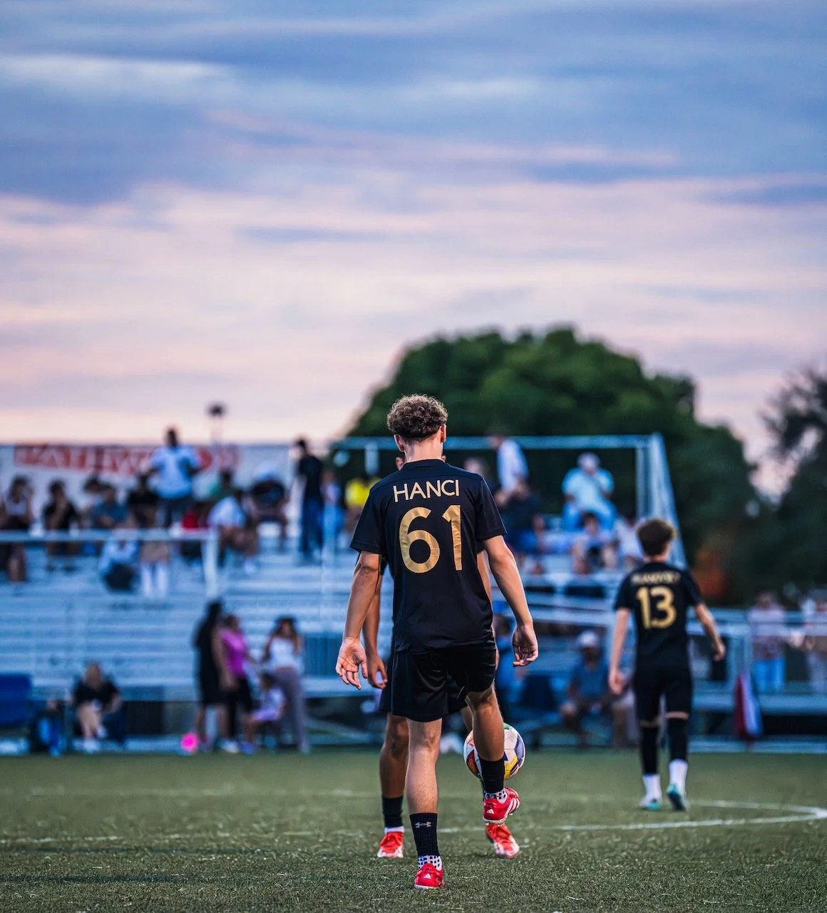 Young soccer player in black jersey number 61 with the name 'Hanci' on the back, standing on the field and preparing to kick a soccer ball during dusk, with fans and seated spectators in the background.