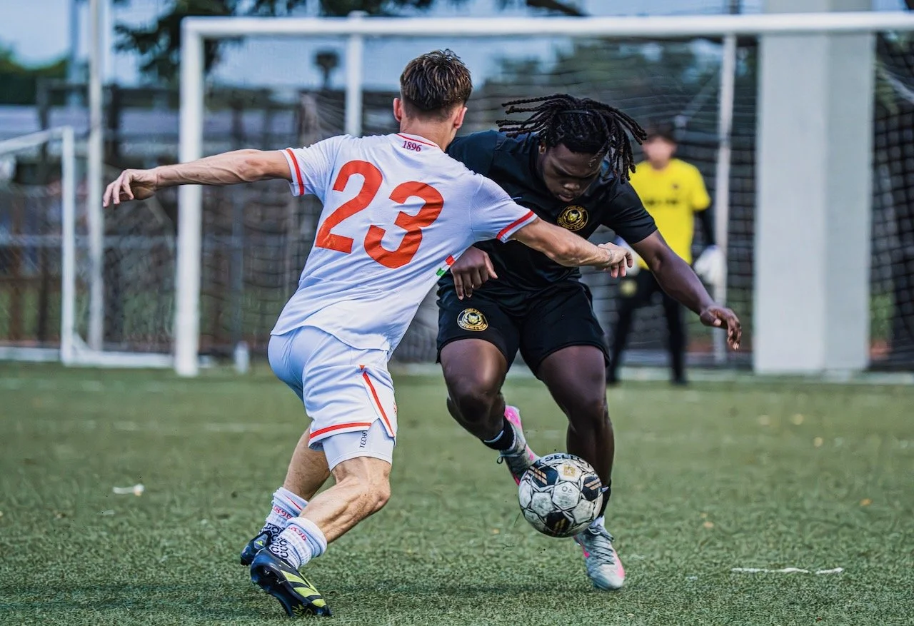 Two soccer players fighting for control of the ball on a field, with a goalkeeper in the background.