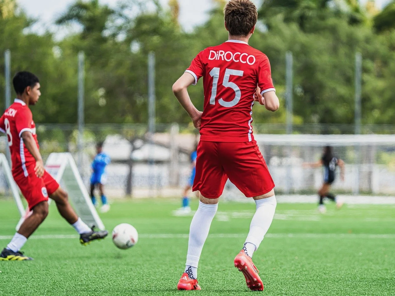 Children playing soccer on a green field, with one kid in a red uniform and the number 15 on the back, and other kids in the background.