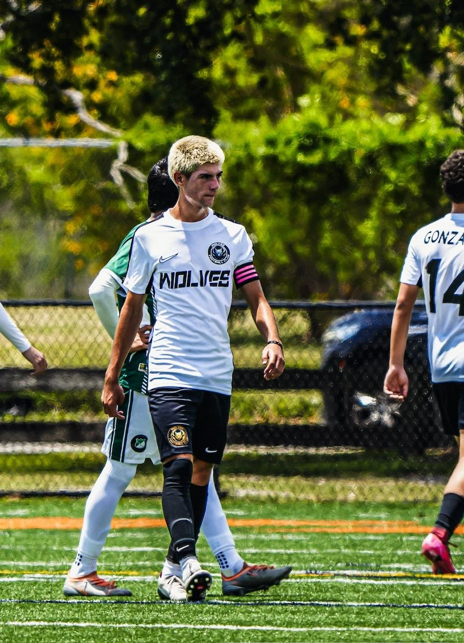 A young male soccer player with bleached blond hair, wearing a white jersey with a black and pink captain's armband, black shorts, and black socks, walks on a green soccer field during daytime. Other players and a chain-link fence are visible in the