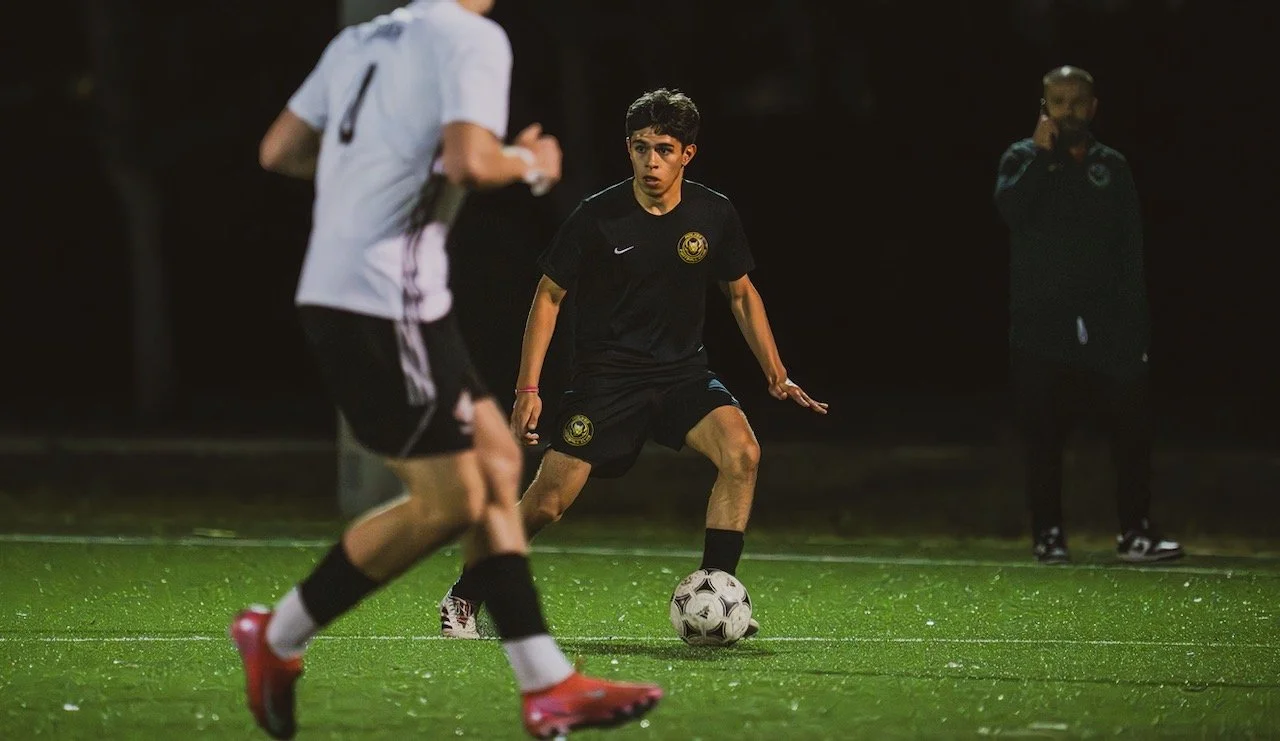 A young soccer player in a black uniform with a crest on the chest controls the ball during a nighttime game on a green field. A player in a white jersey and black shorts runs toward him. Two other individuals are in the background, one on the phone