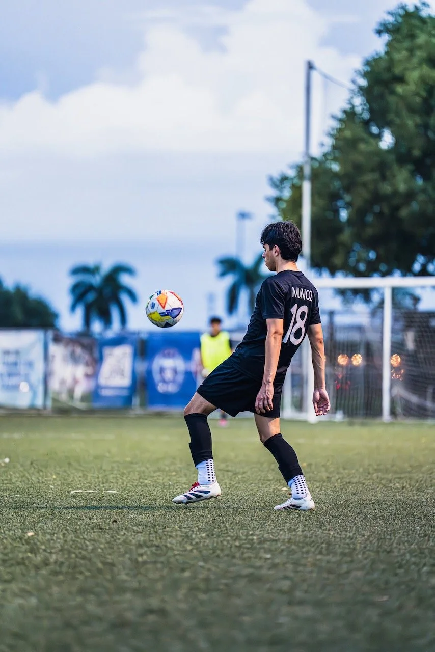 A soccer player in black uniform with number 18 is on a field, preparing to kick a colorful soccer ball during late afternoon or evening, with trees and a goalpost in the background.