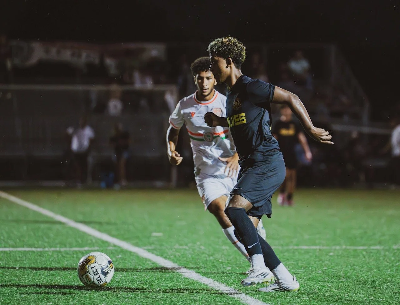 Two soccer players competing for the ball on a green field at night, with spectators in the background.