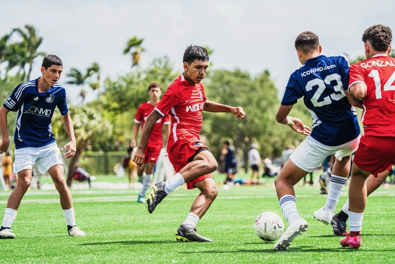Young boys playing soccer on a grass field outdoors during daytime, with some trees and people in the background.