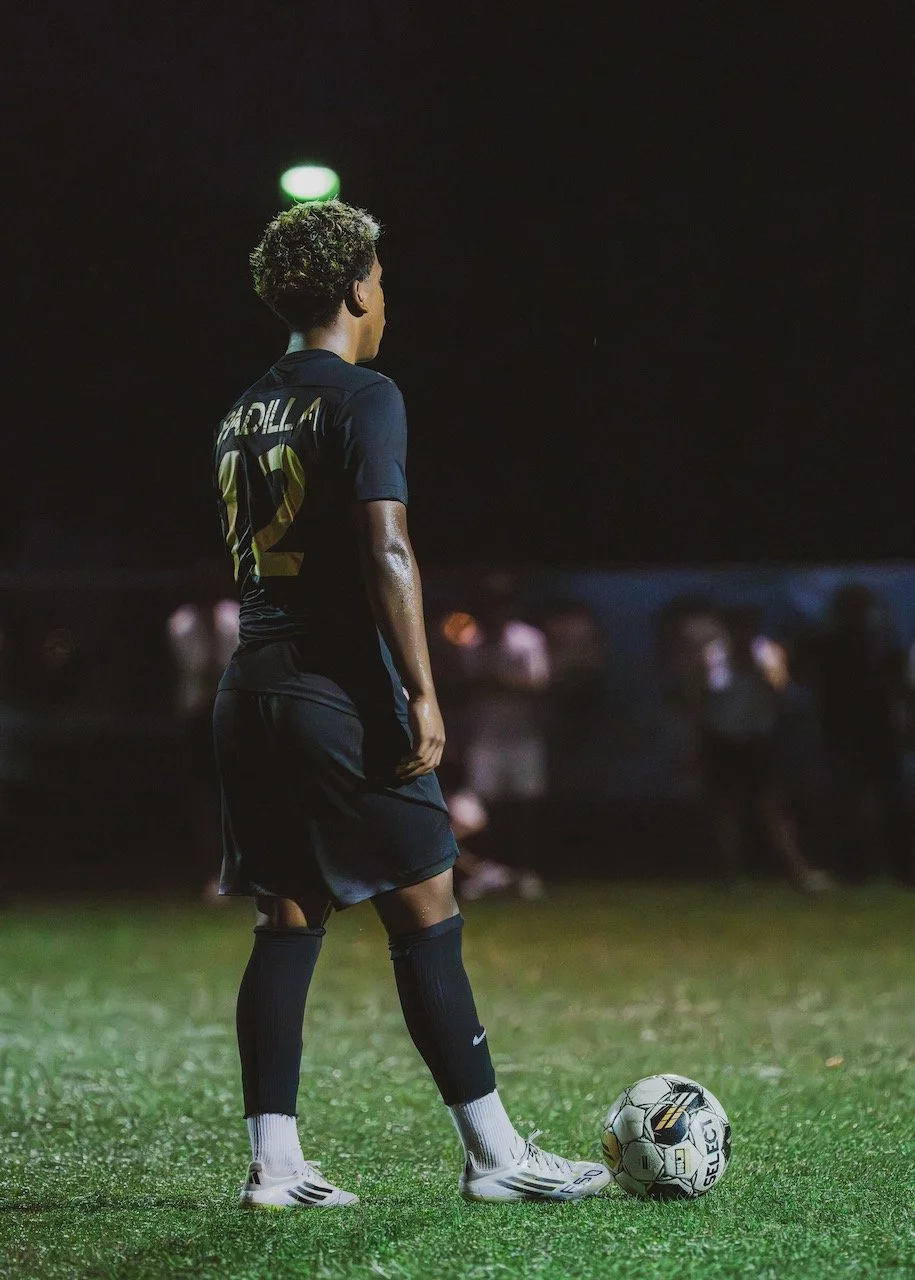 A young soccer player wearing a black uniform with the name Cardella and number 12, standing on a green field at night, preparing to kick a soccer ball.