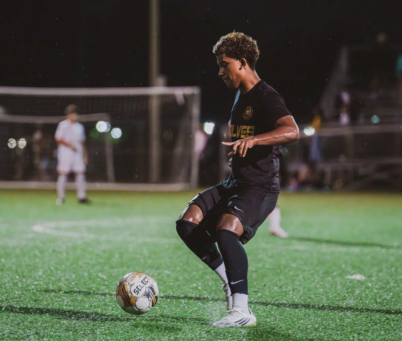 A soccer player in a black uniform is about to kick a soccer ball on a field during night play, with two blurred figures in the background and rain visible.