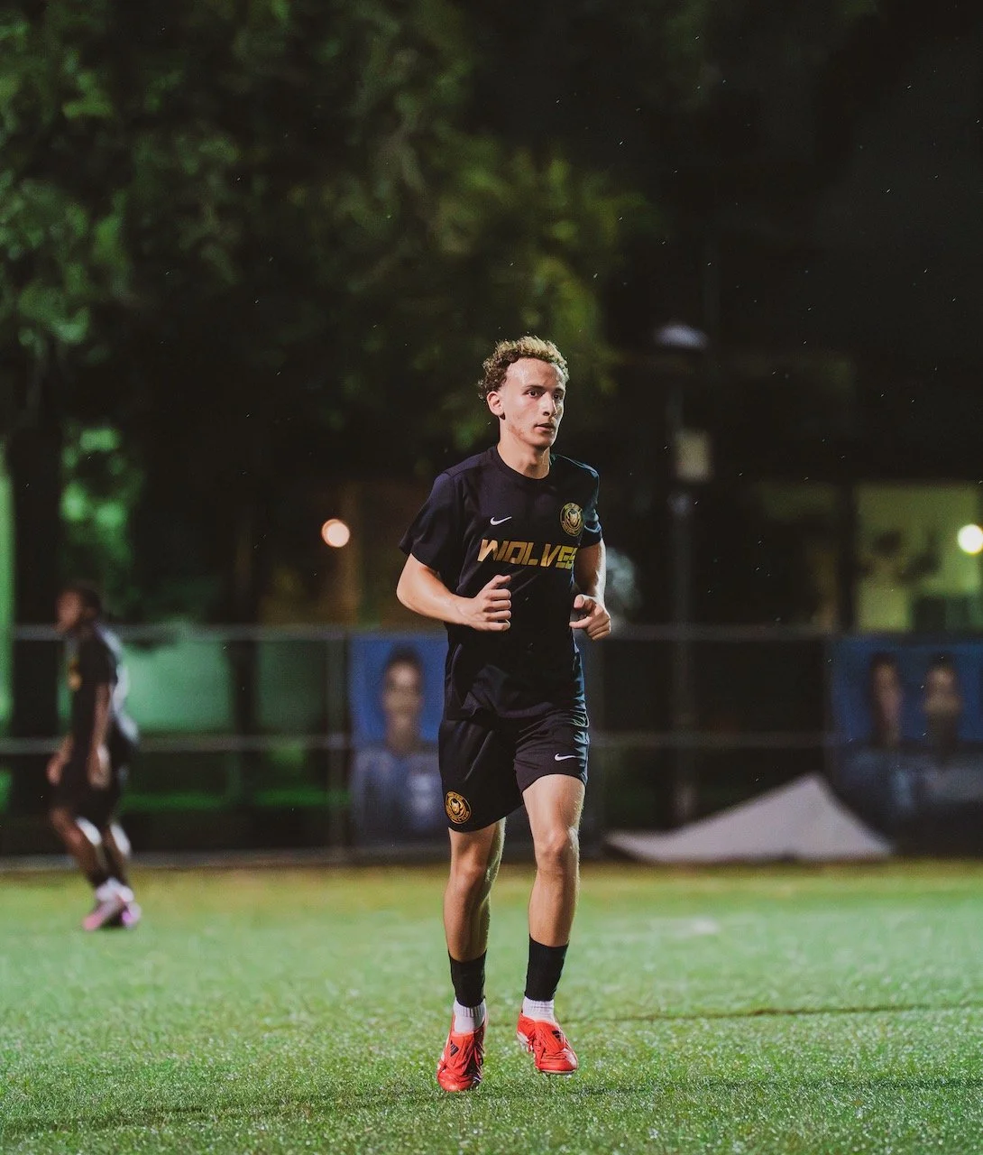 A young male soccer player practicing on a field at night, wearing a black uniform with gold accents and red cleats.