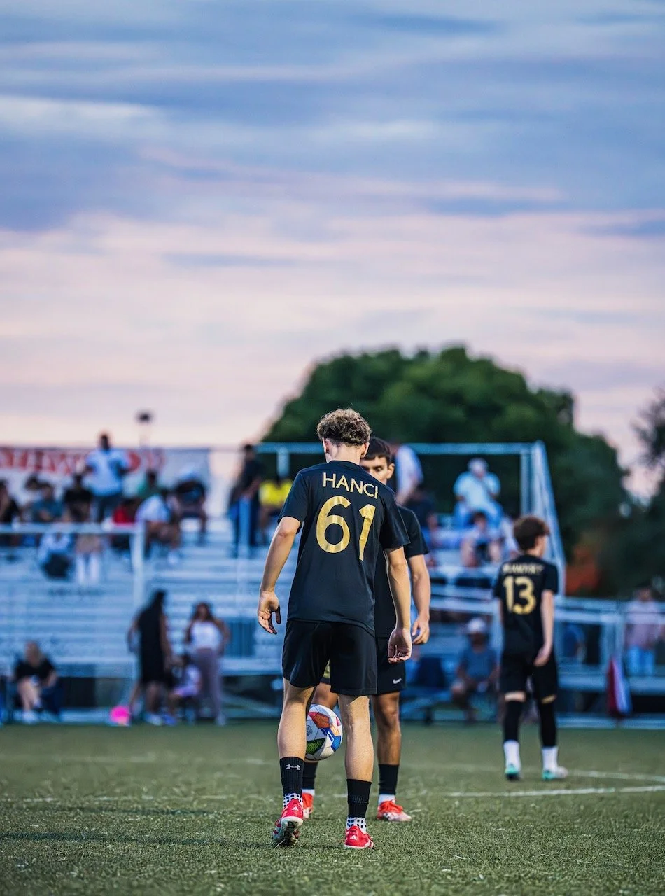 Young soccer players on a field during a game, with one player holding a soccer ball, spectators sitting on bleachers in the background, and a partly cloudy sky overhead.