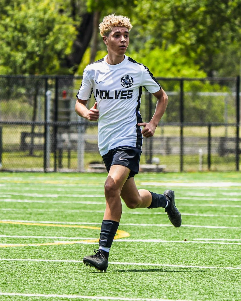 A young male soccer player running on a green field wearing a white jersey with black accents and the word 'WOLVES' on it, black shorts, black cleats, and black socks.