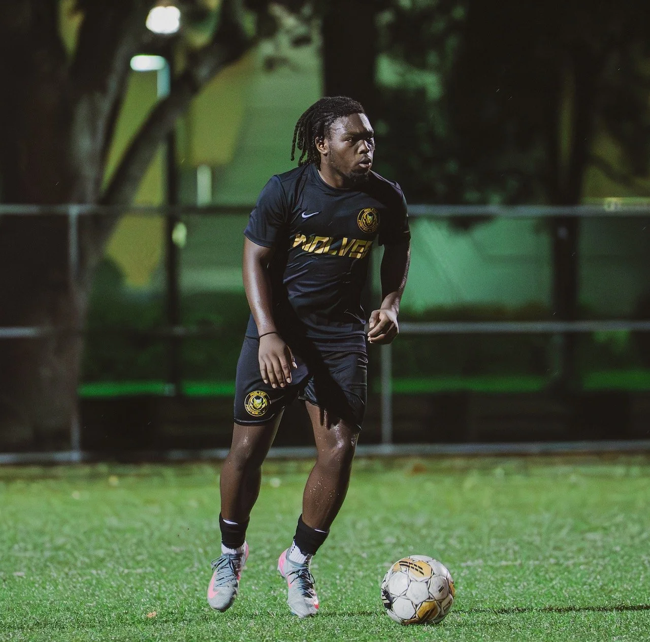 A male soccer player with dreadlocks wearing a black uniform with yellow accents is dribbling a soccer ball on a grass field during evening or night under artificial lights.