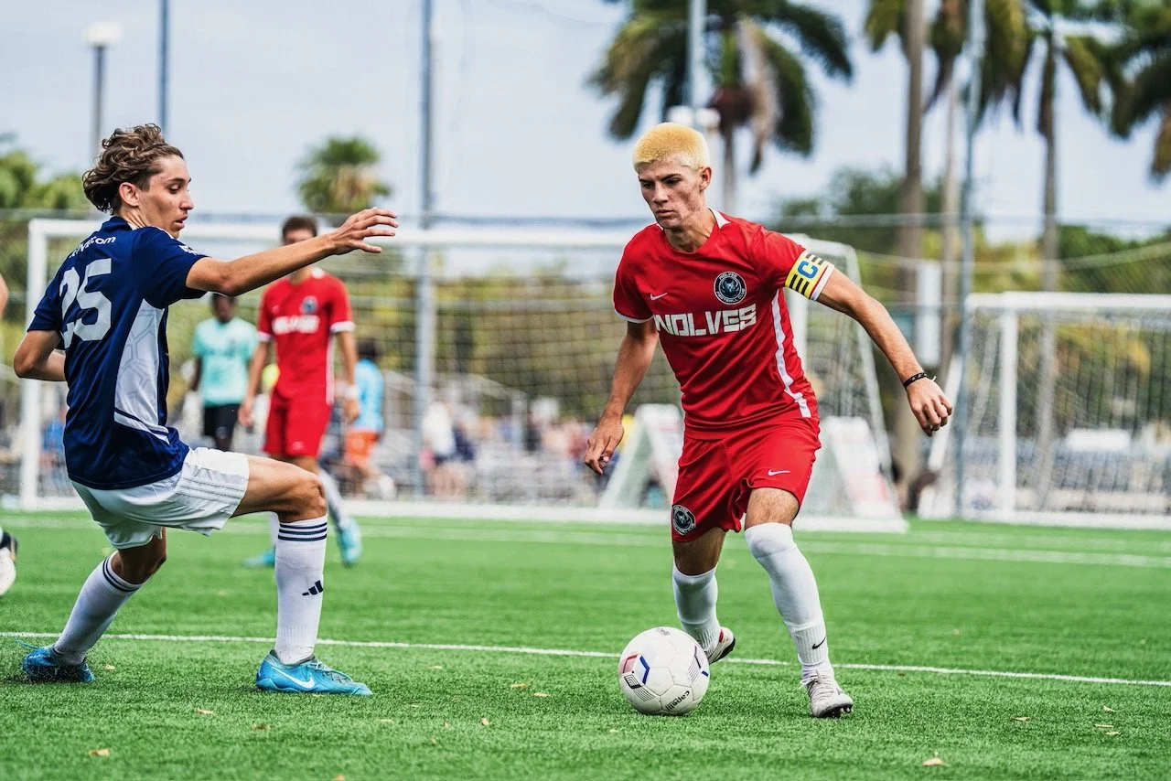 A soccer match with two female players competing for the ball on a green field. One player in a red uniform with blonde hair is dribbling the ball, while the other in a blue uniform with brown hair tries to block her.